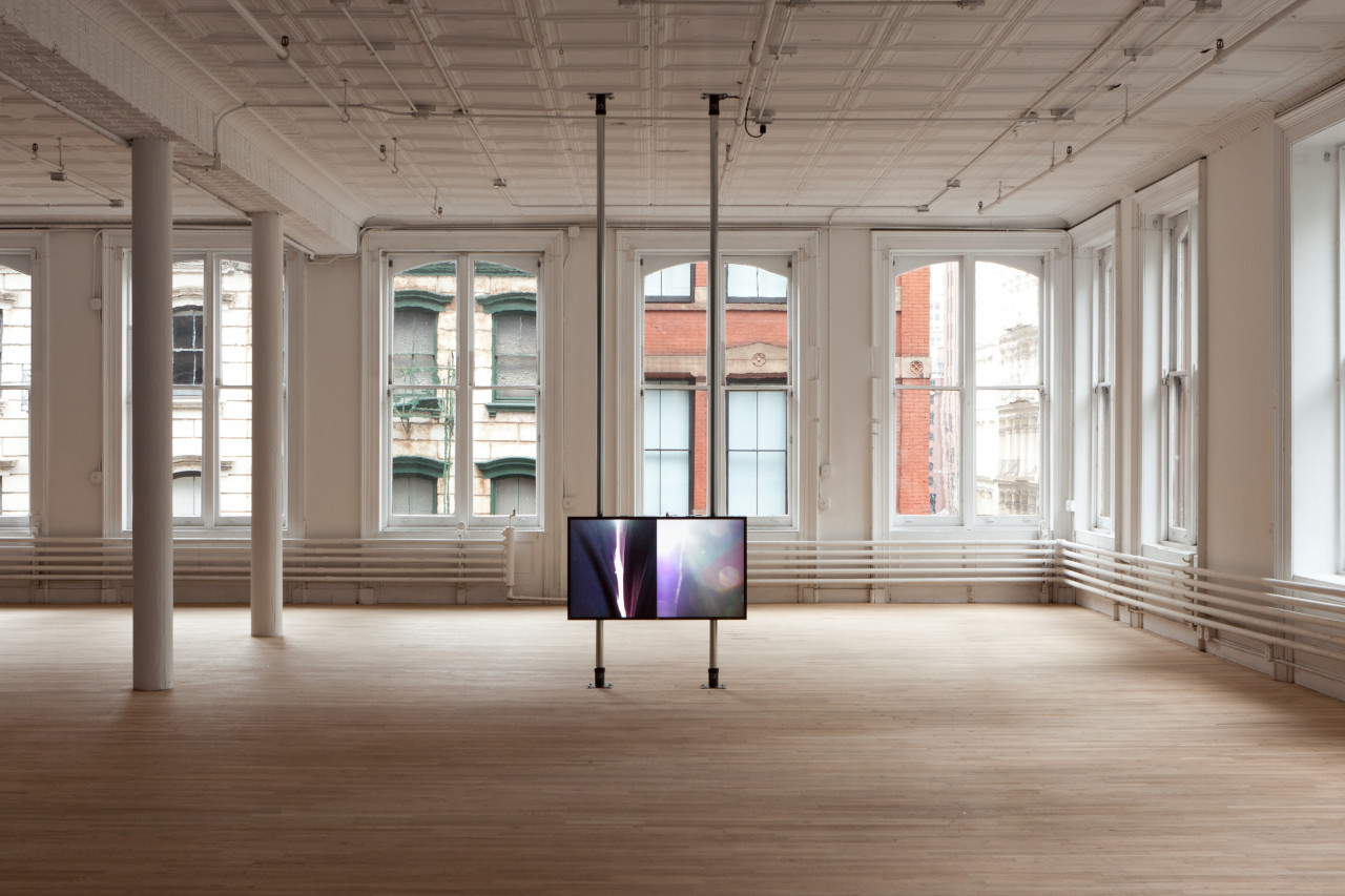 A single screen showing a splitscreen image is mounted on two poles that stretch floor to ceiling. The screen stands alone in a large, open space with windows in the background.