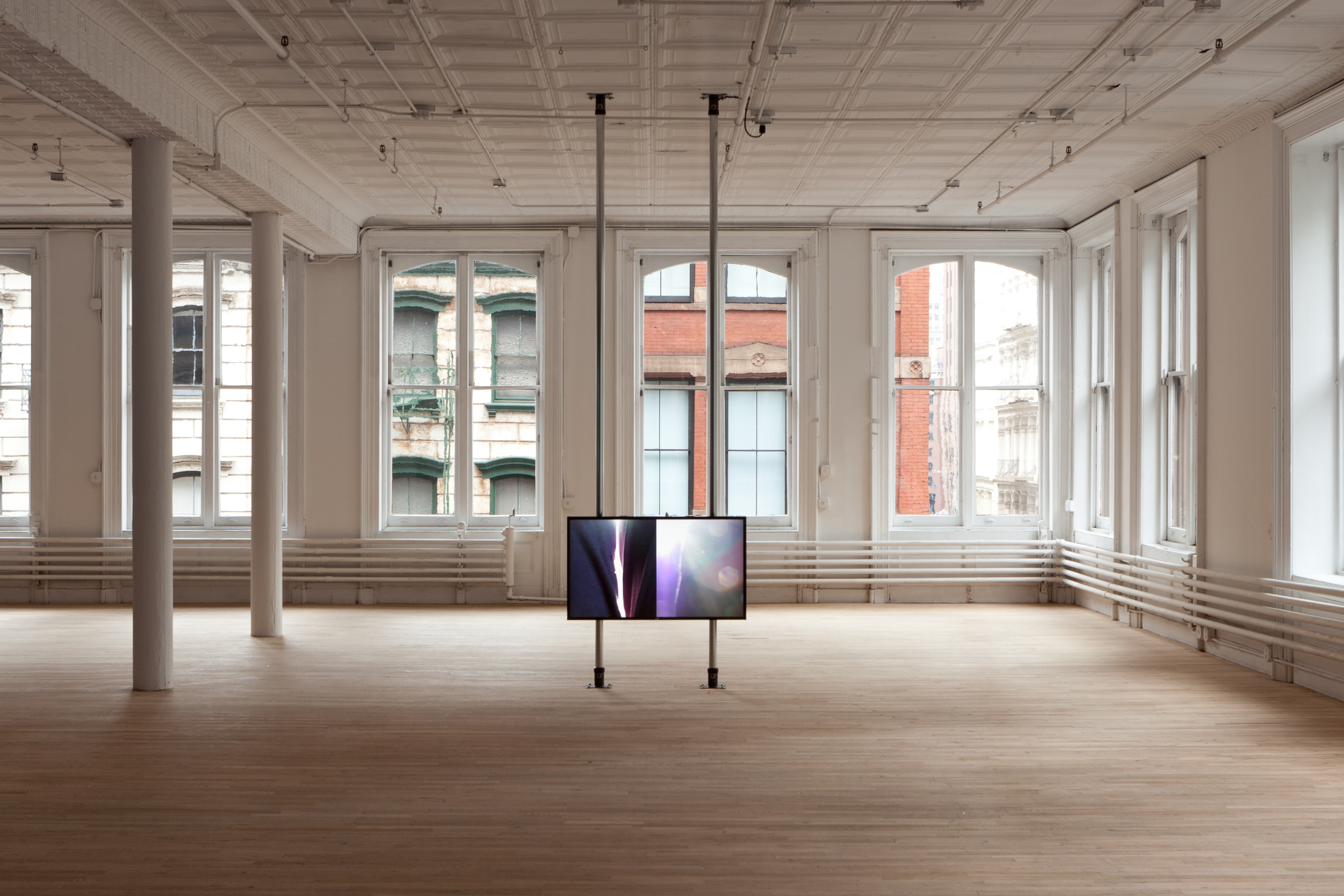 A single screen showing a splitscreen image is mounted on two poles that stretch floor to ceiling. The screen stands alone in a large, open space with windows in the background.
