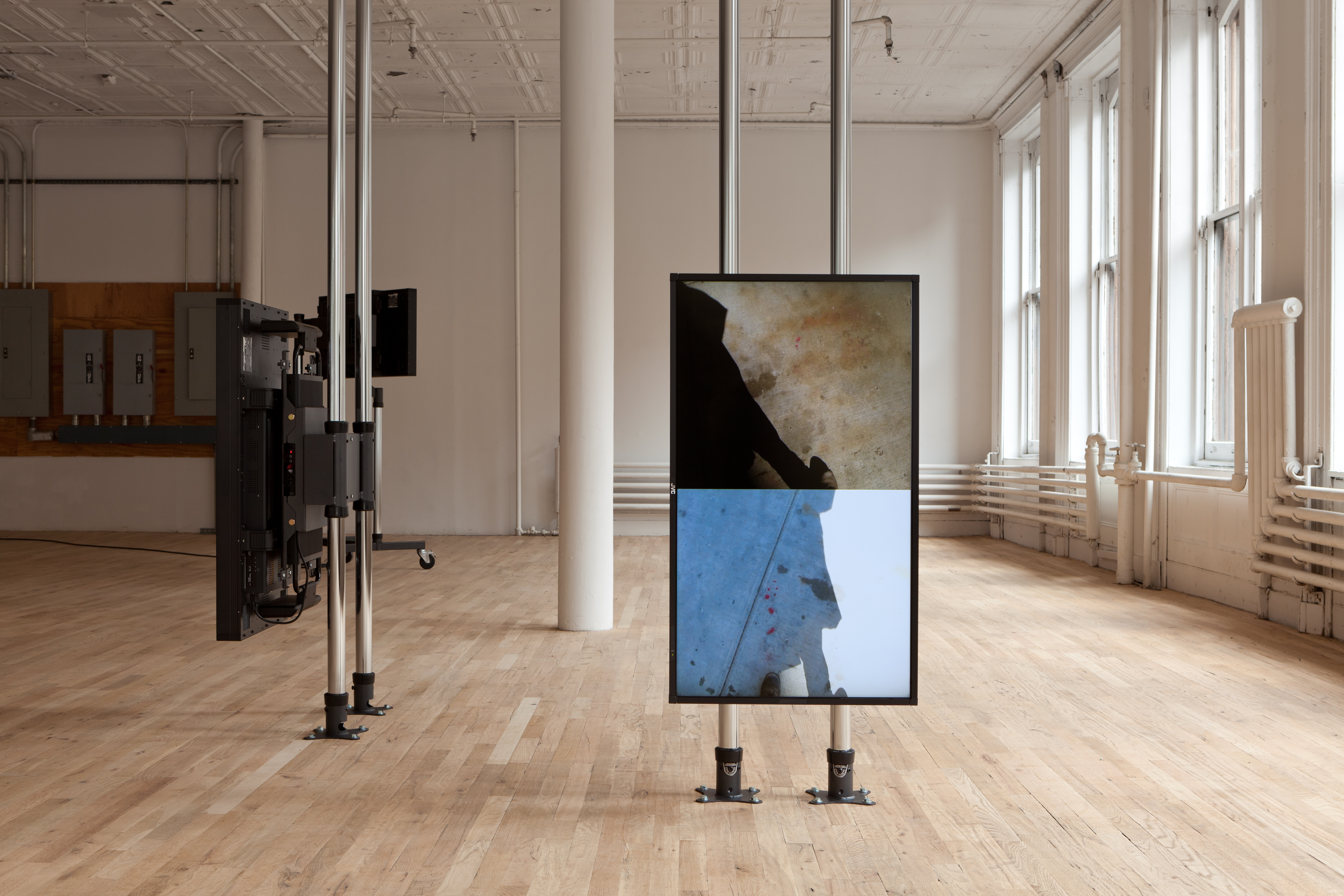 A portrait-oriented screen is mounted on two metal floor to ceiling poles. The screen is split to show two different images of shadows on the ground. At a 90-degree angle facing left, another screen is mounted on two metal floor to ceiling poles. A third screen is visible in the background, facing away, mounted on two metal poles atop a wheeled base.