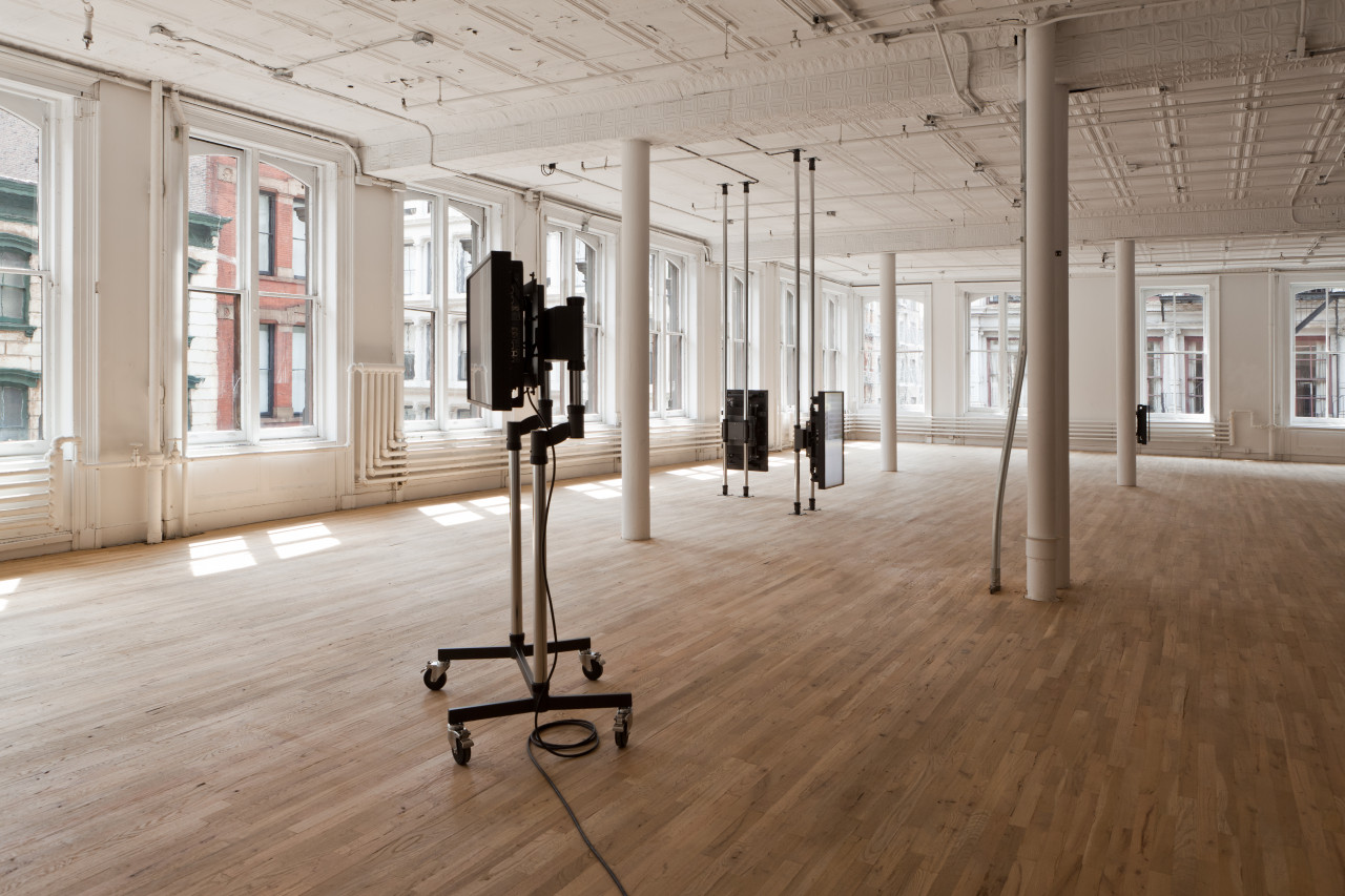 A wide shot of a gallery space with wood floors and large windows. In the foreground is a screen mounted on two metal poles on a wheeled base. In the midground, there are two screens mounted on floor to ceiling metal poles arranged at a 90-degree angle to each other. In the background, there is another screen just visible from behind a white column.