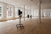 A wide shot of a gallery space with wood floors and large windows. In the foreground is a screen mounted on two metal poles on a wheeled base. In the midground, there are two screens mounted on floor to ceiling metal poles arranged at a 90-degree angle to each other. In the background, there is another screen just visible from behind a white column.