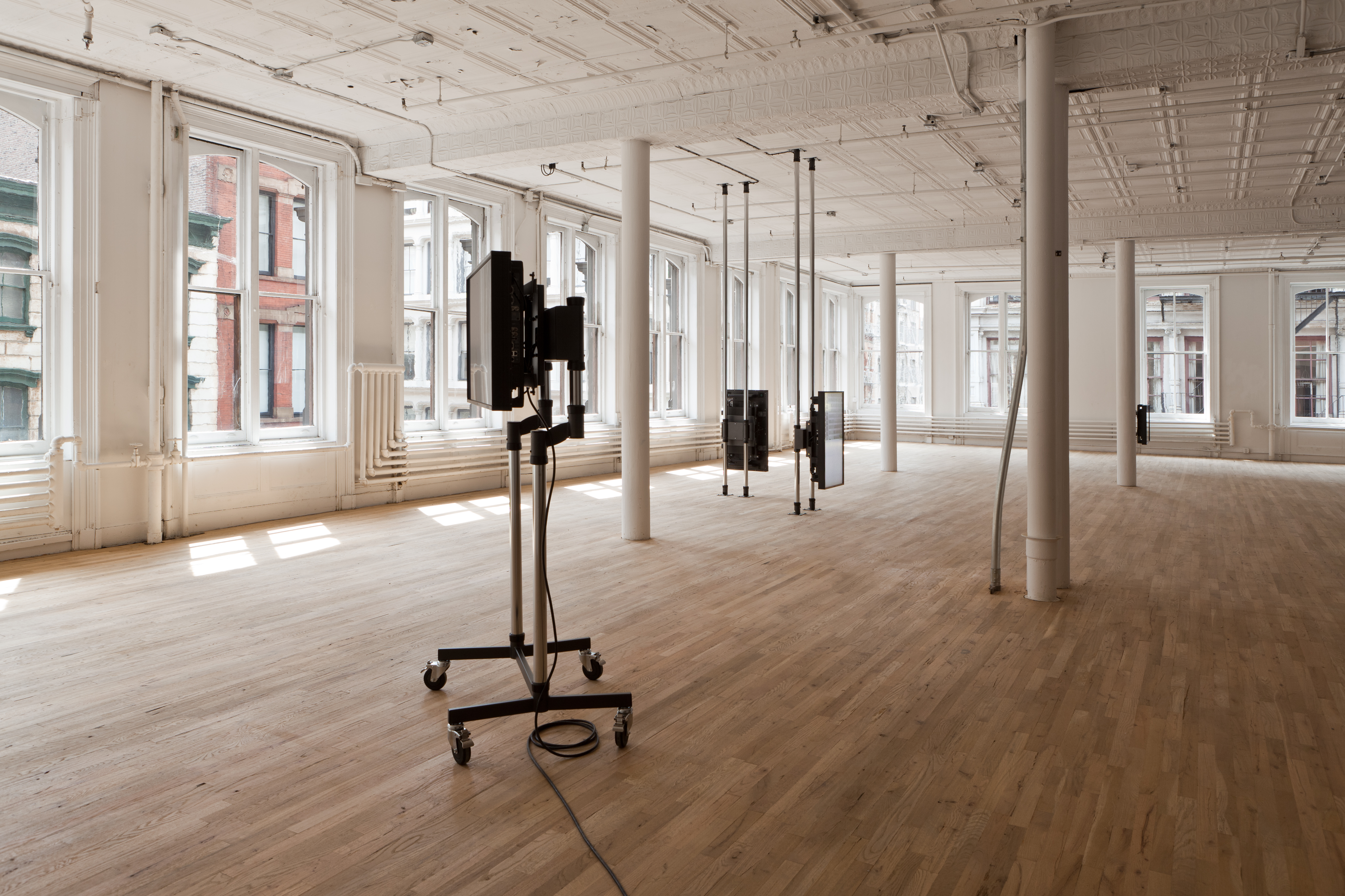 A wide shot of a gallery space with wood floors and large windows. In the foreground is a screen mounted on two metal poles on a wheeled base. In the midground, there are two screens mounted on floor to ceiling metal poles arranged at a 90-degree angle to each other. In the background, there is another screen just visible from behind a white column.