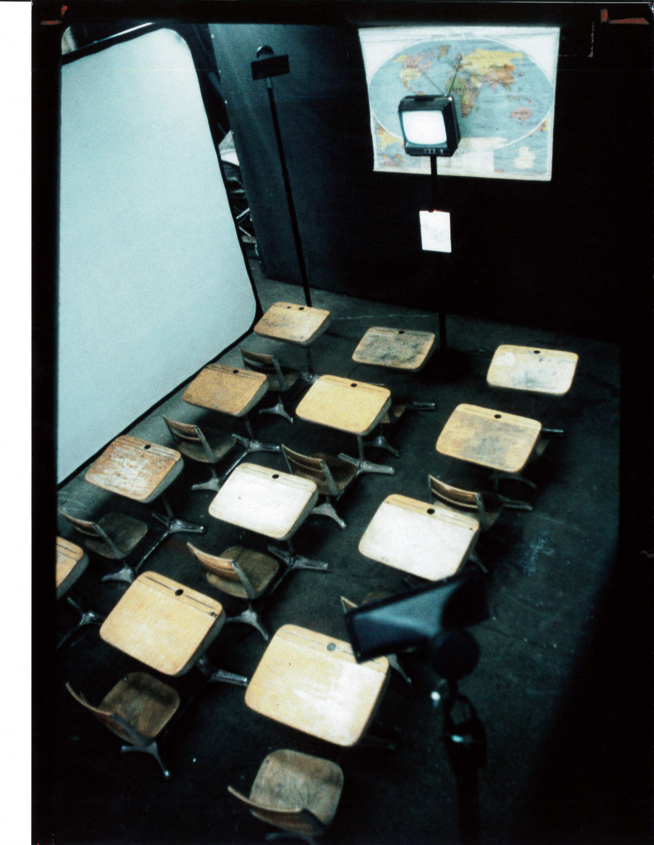 A photograph, taken from above, of rows of school desks facing a television with a map behind it.