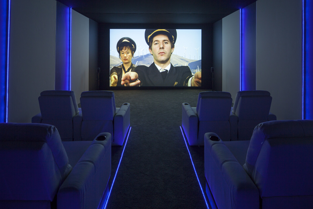 A view down an isle, between theater seating and blue track lighting, to a projected video screen. On the back wall, a still image of a man, in a pilots uniform, gestures toward the viewers with two fingers. Hito Stereyl stands behind him with her arm outstretched.