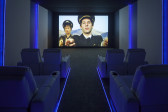 A view down an isle, between theater seating and blue track lighting, to a projected video screen. On the back wall, a still image of a man, in a pilots uniform, gestures toward the viewers with two fingers. Hito Stereyl stands behind him with her arm outstretched.