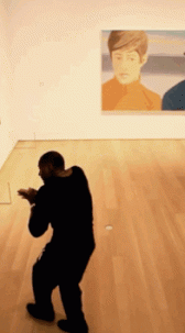 A moving image of a person, dressed in black, side stepping slowly in the gallery as the camera pans over to them.