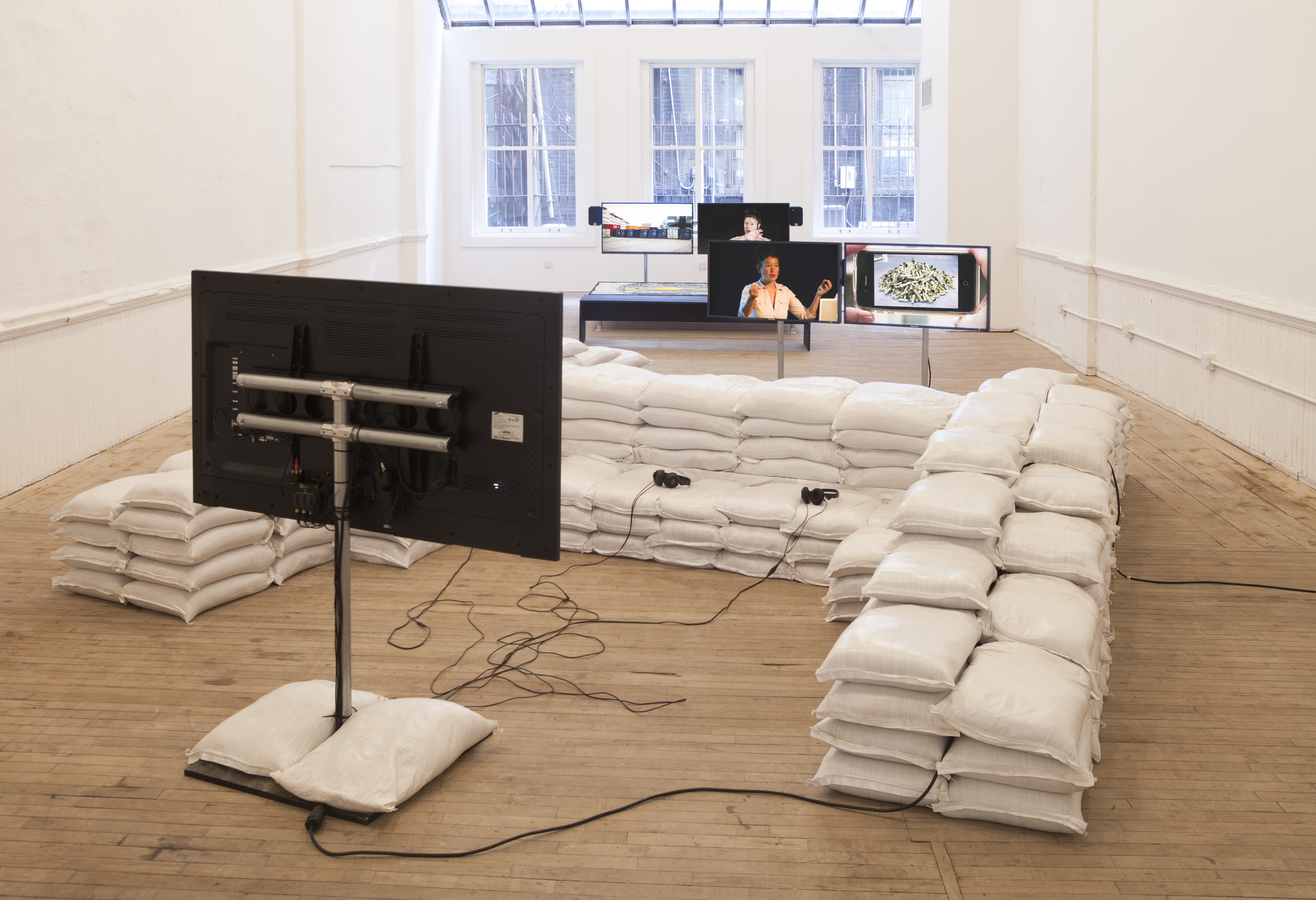 A monitor facing a seating arrangement constructed of stacked pillows. Two pairs of monitors are suspended on poles in the background. An image of Hito Steyerl is seen in both pairs; next to an image of a pile of cigarettes and an image of shipping containers.