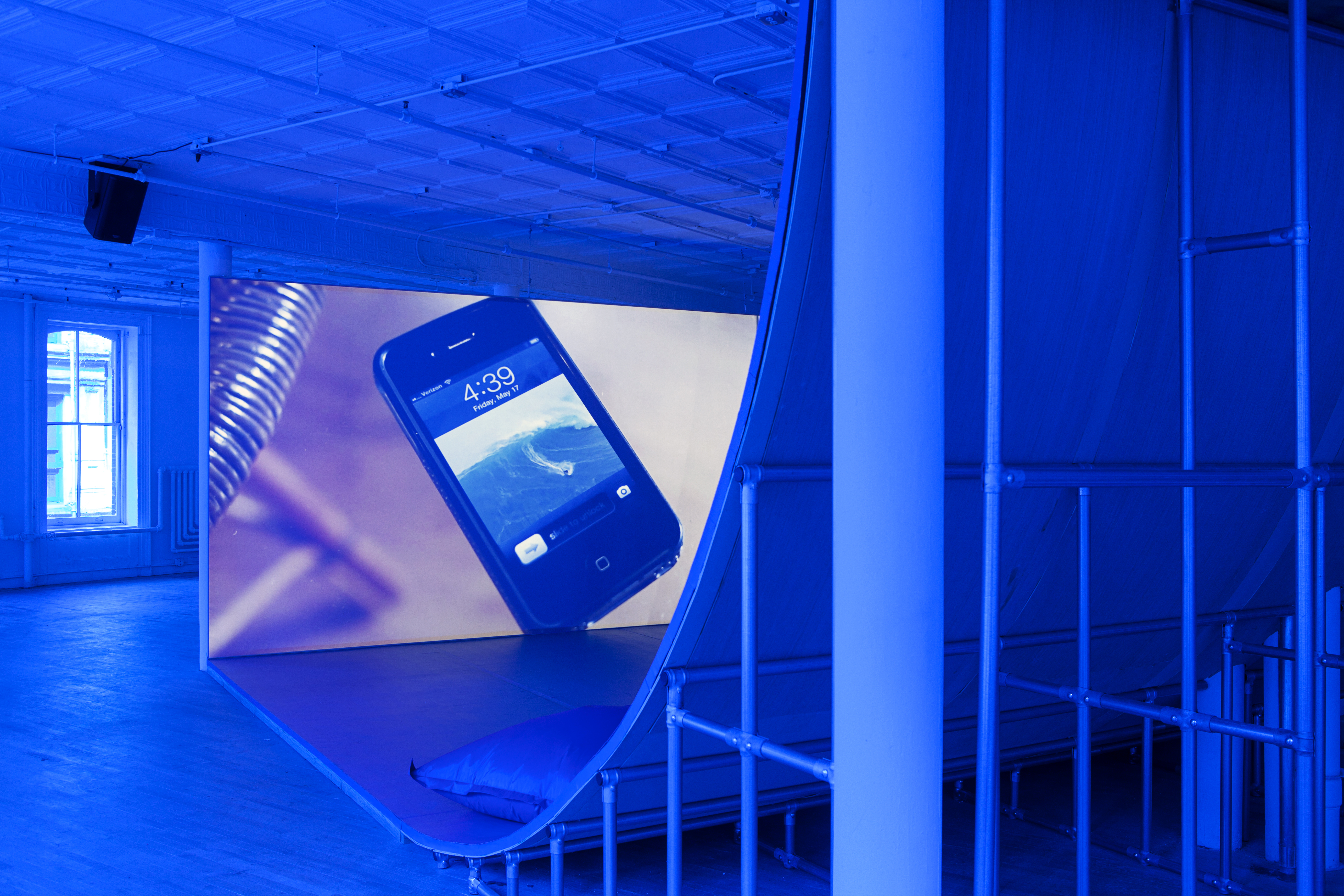 An angled view of a large ramp leading to a large video monitor screen. The room is tinted blue. On the screen, an iPhone 4 sits on a glass table, it's lock screen on view; the time reads, 