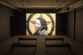 A dim room with four benches in front of a large video screen. A black and white image of a woman in front of a red star and a wreath is projected.