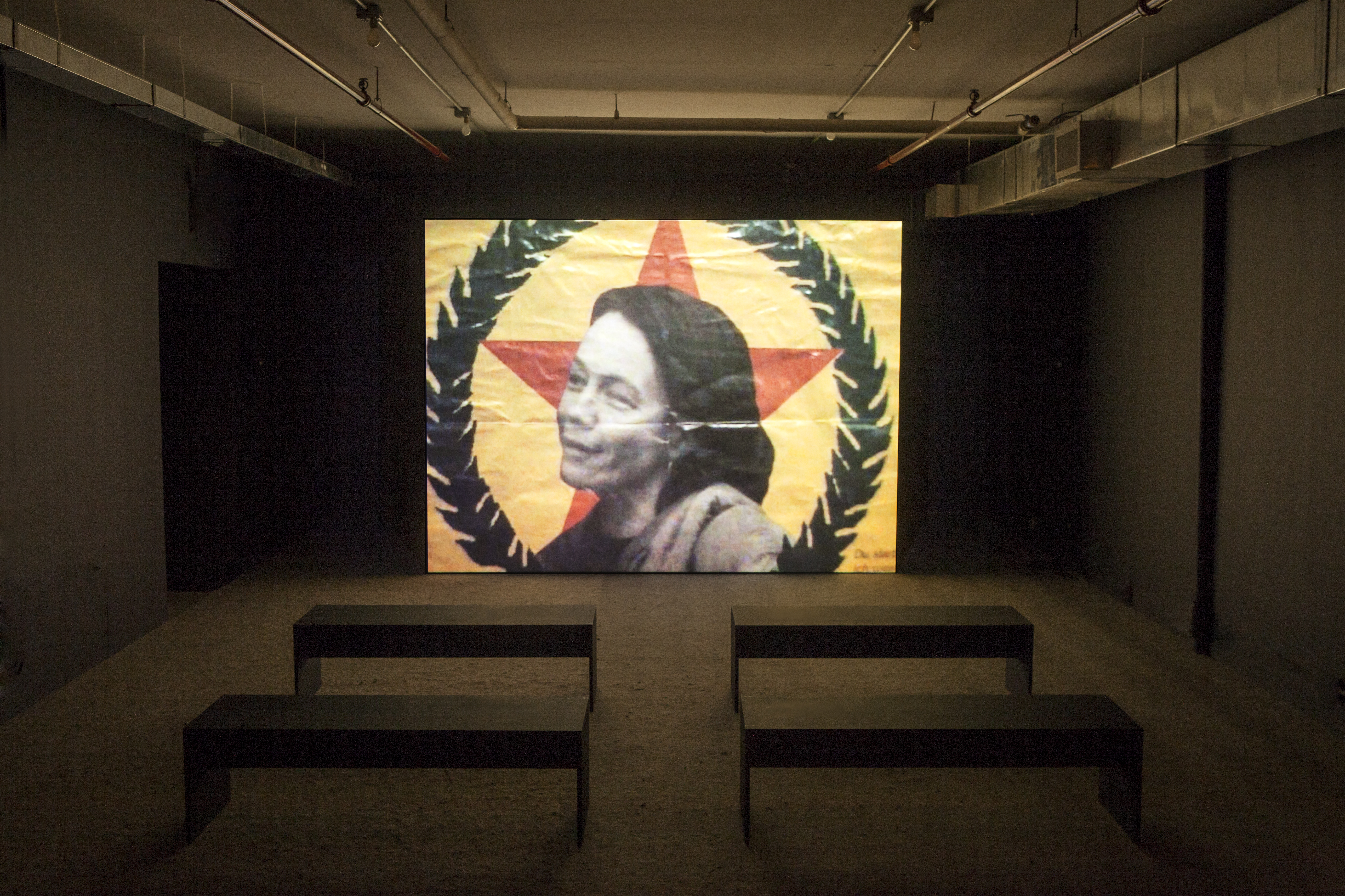 A dim room with four benches in front of a large video screen. A black and white image of a woman in front of a red star and a wreath is projected.