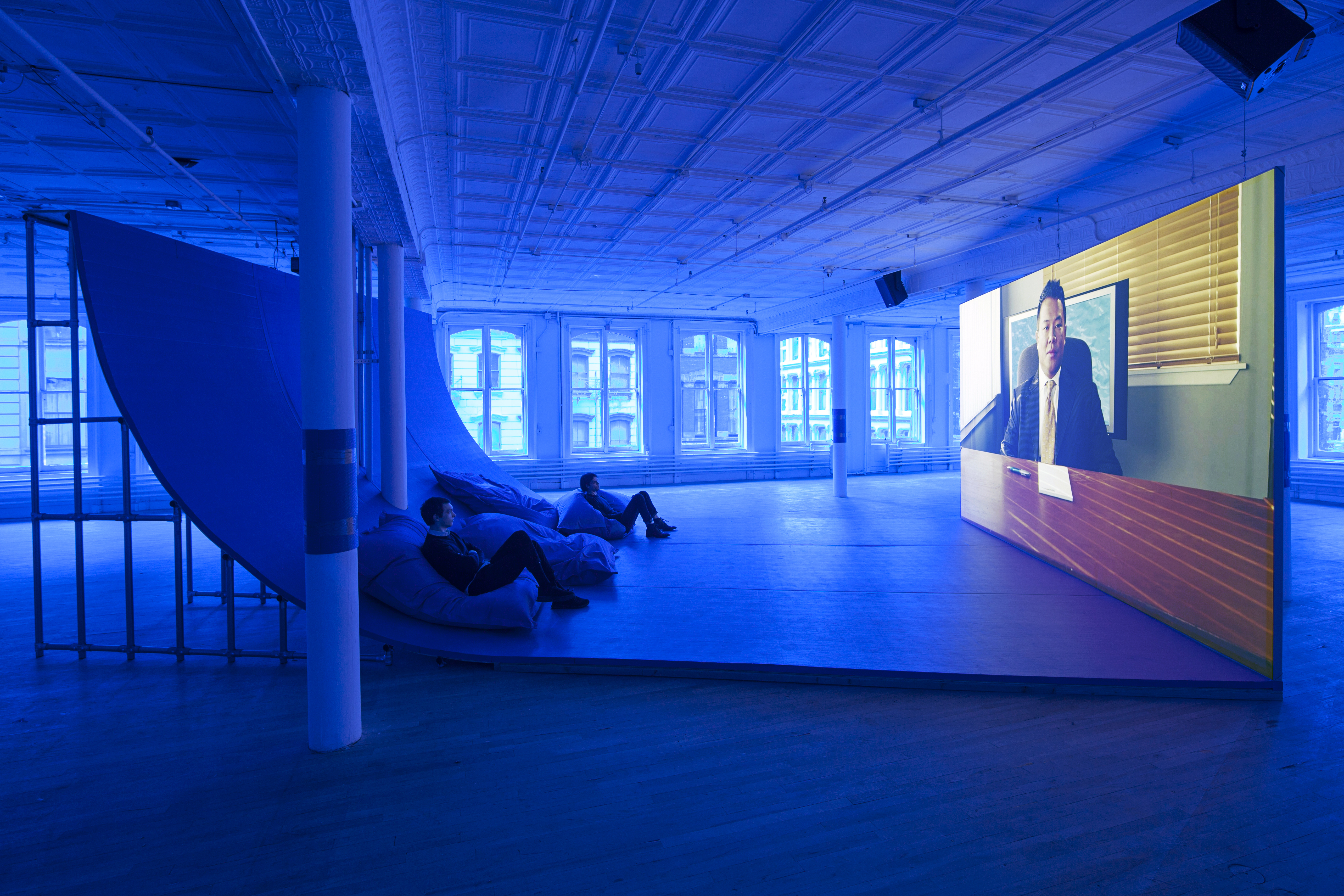 A side view of a large video monitor screen, sitting on the ground, with a large ramp extending from the base. Two viewers sit on large pillows at the curve of the ramp. The room is tinted blue. On the screen, there is a man in a sitting in an office with a note pad and a pen at the table in front of him.