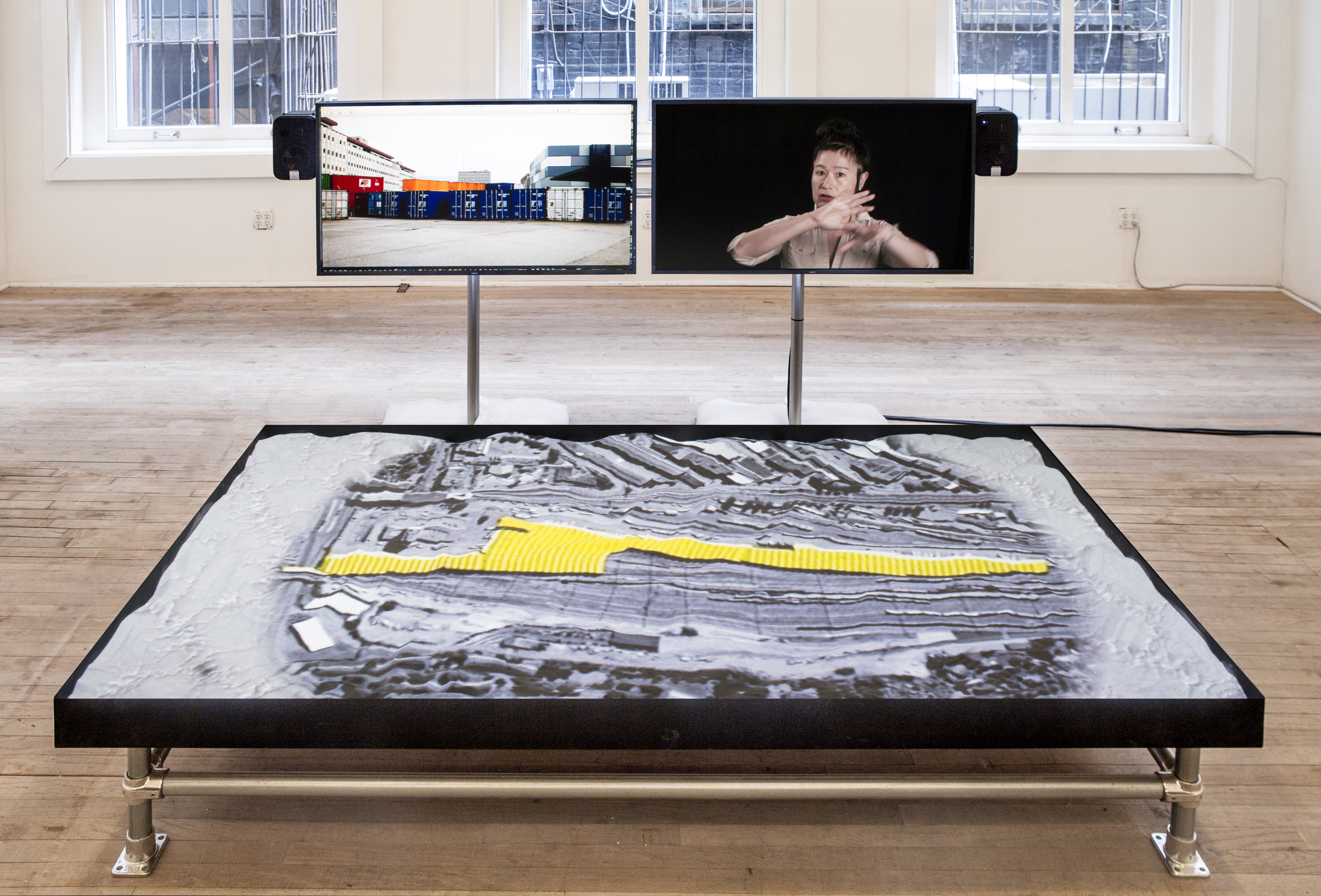 One large monitor lays horizontally on a low frame in front of two smaller monitors suspended on poles. The bottom monitor projects a black-and-white aerial view of a building that is highlighted yellow. The left monitor at eye level displays shipping containers. The right monitor shows Hito Steyerl in front of a black background.