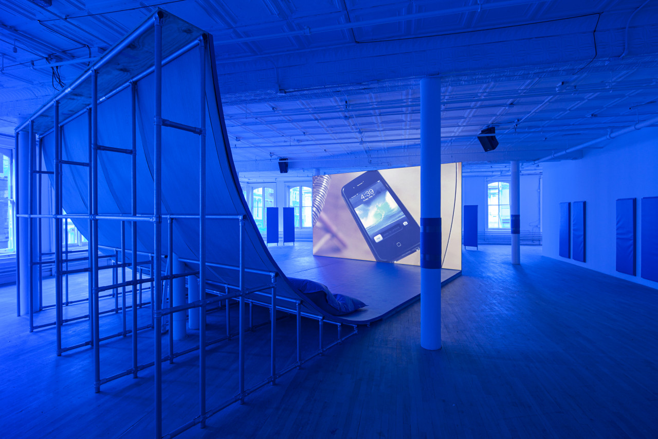 An angled view of a large ramp looking on to a large video monitor screen positioned at the base. The room is tinted blue. On the screen, an iPhone 4 sits on a glass table, its lock screen on view; the time reads, "4:39."