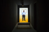 A portrait oriented video is projected in a dark narrow space. A bench sits in front of the video. An image-still of a man, hands clasped at his waist as he faces the viewer, is on the back wall.