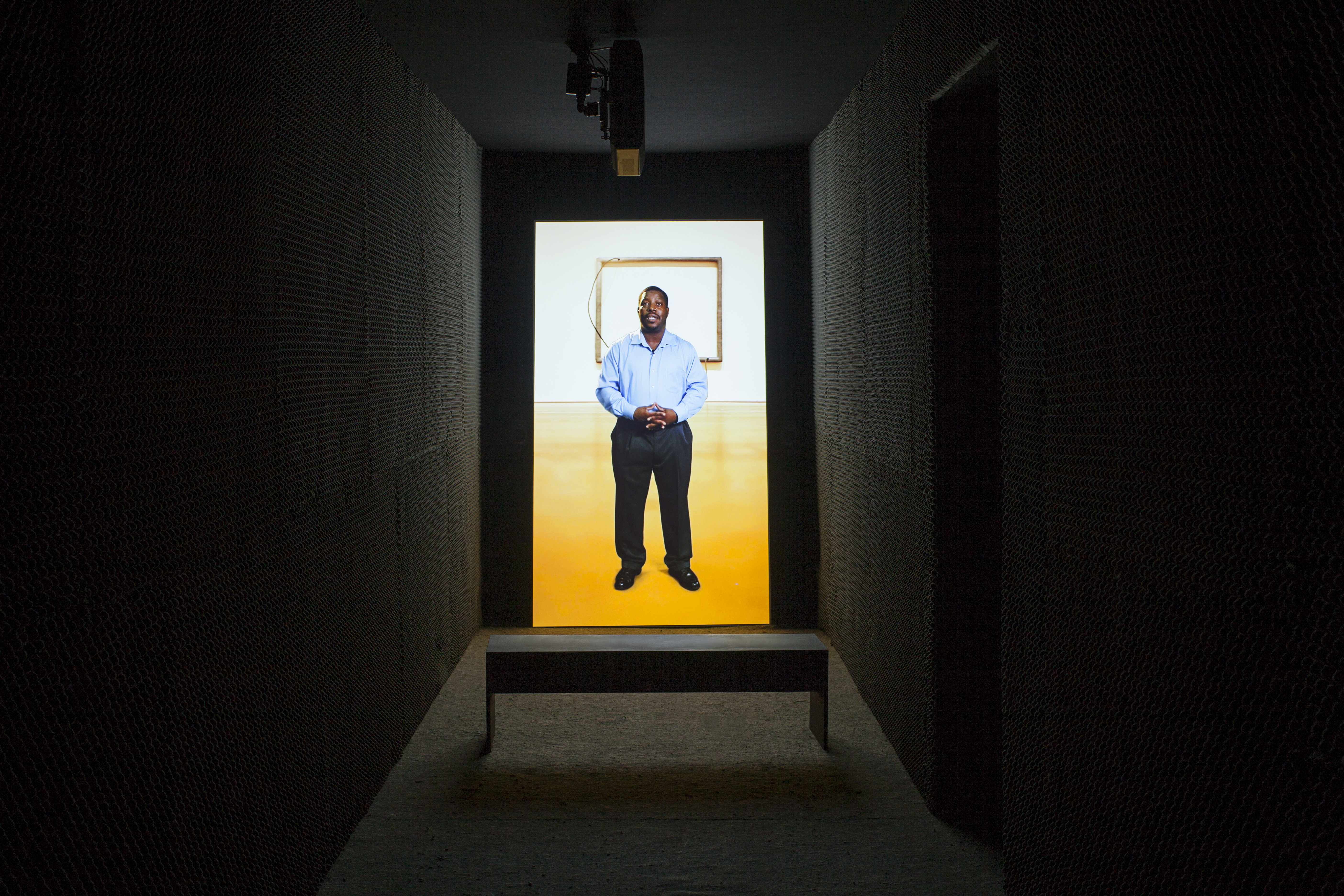 A portrait oriented video is projected in a dark narrow space. A bench sits in front of the video. An image-still of a man, hands clasped at his waist as he faces the viewer, is on the back wall.