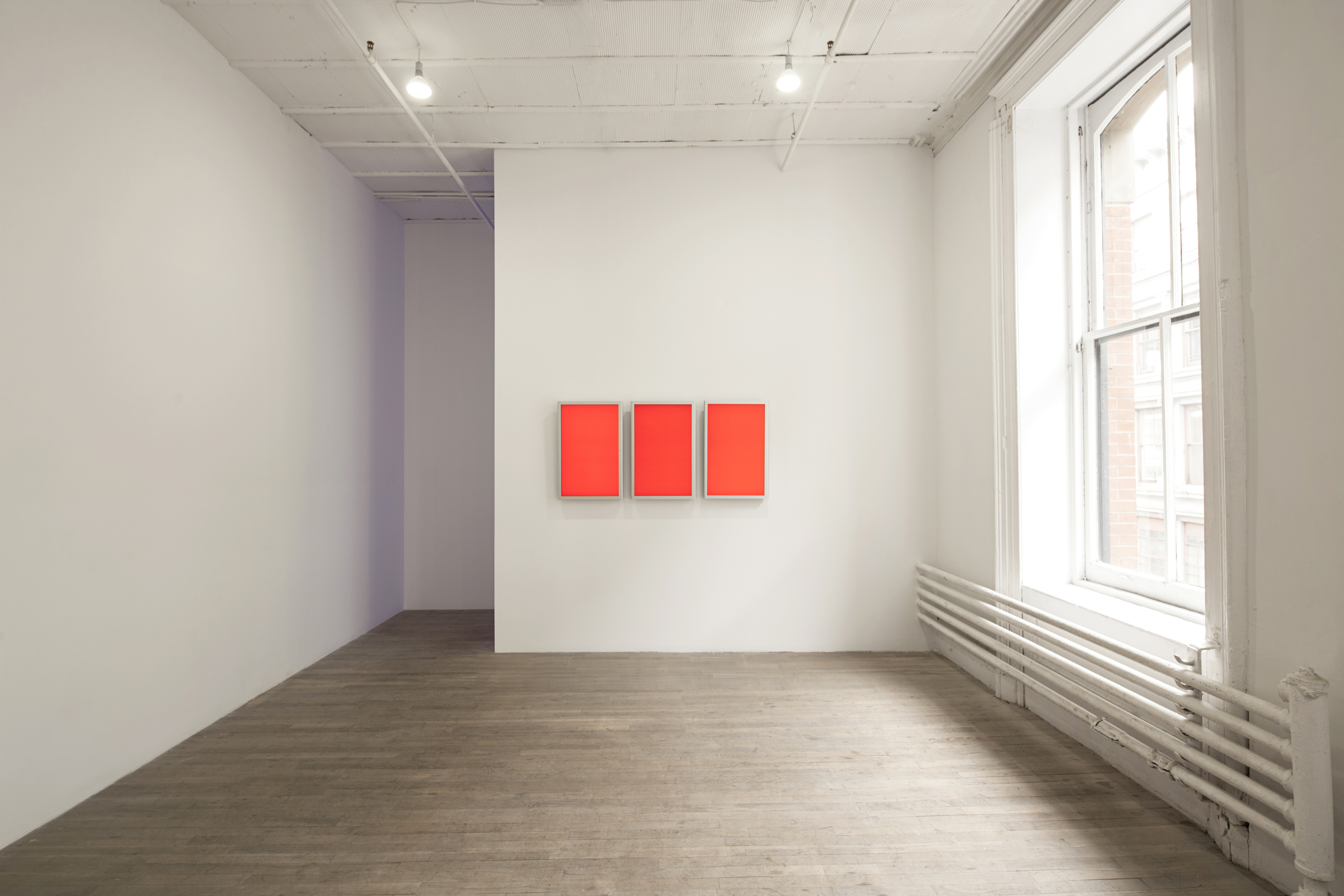 Three red-orange panels in white frames hang on a white wall in an open space.