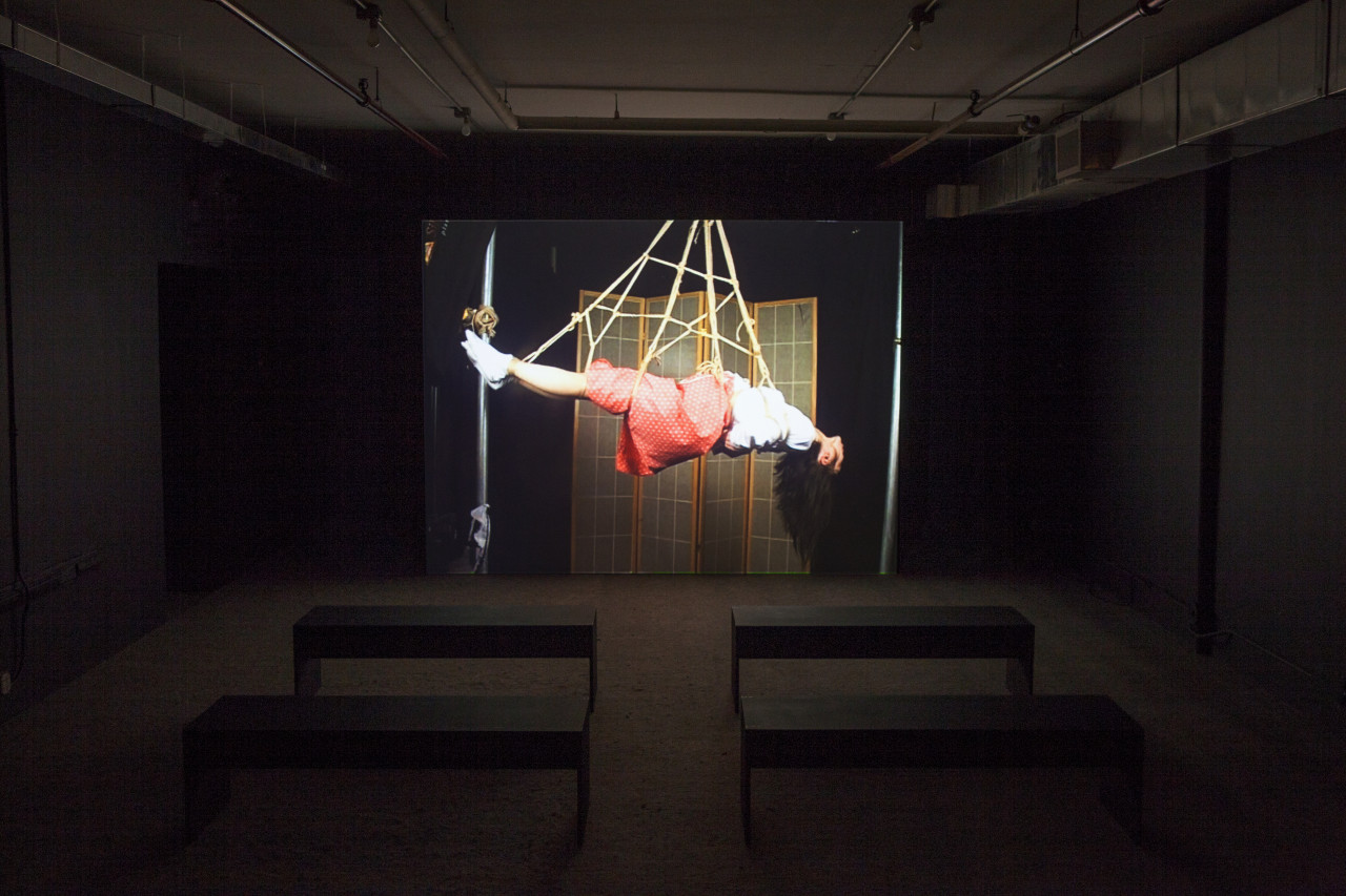 A dim room with four benches in front of a large video projection screen. A still image of a woman suspended by rope is projected.