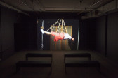 A dim room with four benches in front of a large video projection screen. A still image of a woman suspended by rope is projected.