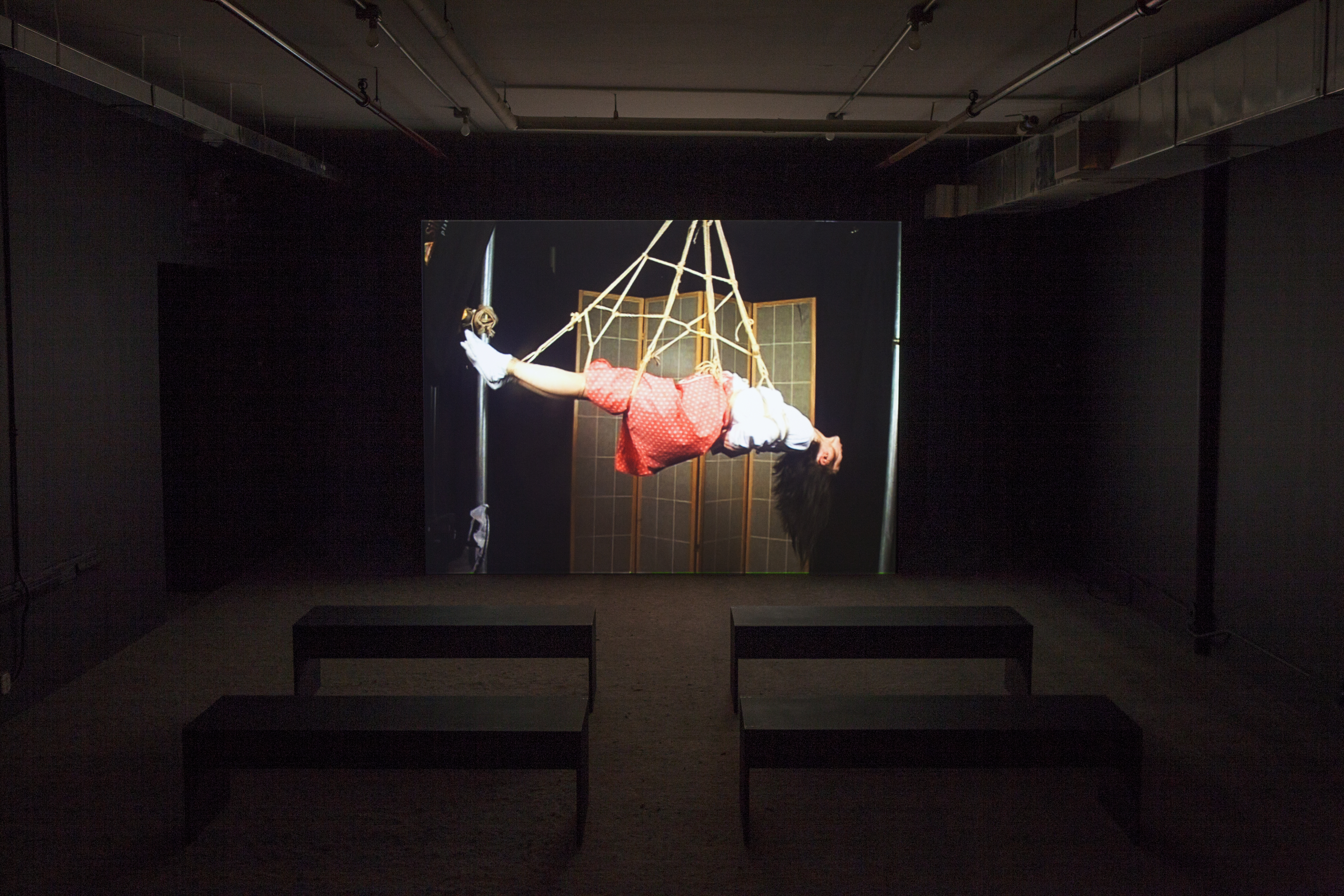 A dim room with four benches in front of a large video projection screen. A still image of a woman suspended by rope is projected.