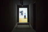 A portrait oriented video is projected in a dark narrow space. A bench sits in front of the video. An image-still of a man, standing next to a frame on a wall that contains a seperate video of a man in a street, is on the back wall.