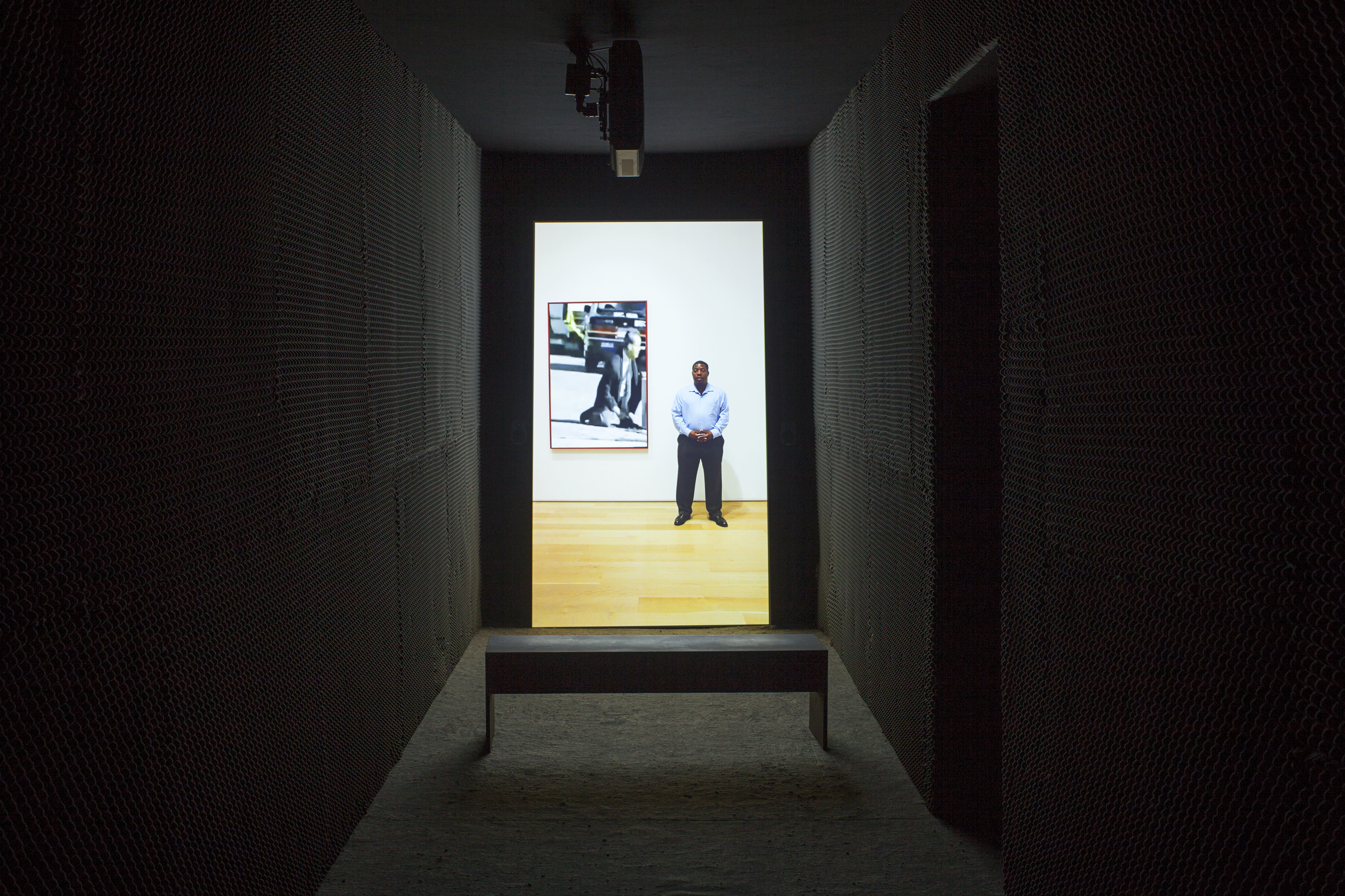 A portrait oriented video is projected in a dark narrow space. A bench sits in front of the video. An image-still of a man, standing next to a frame on a wall that contains a seperate video of a man in a street, is on the back wall.
