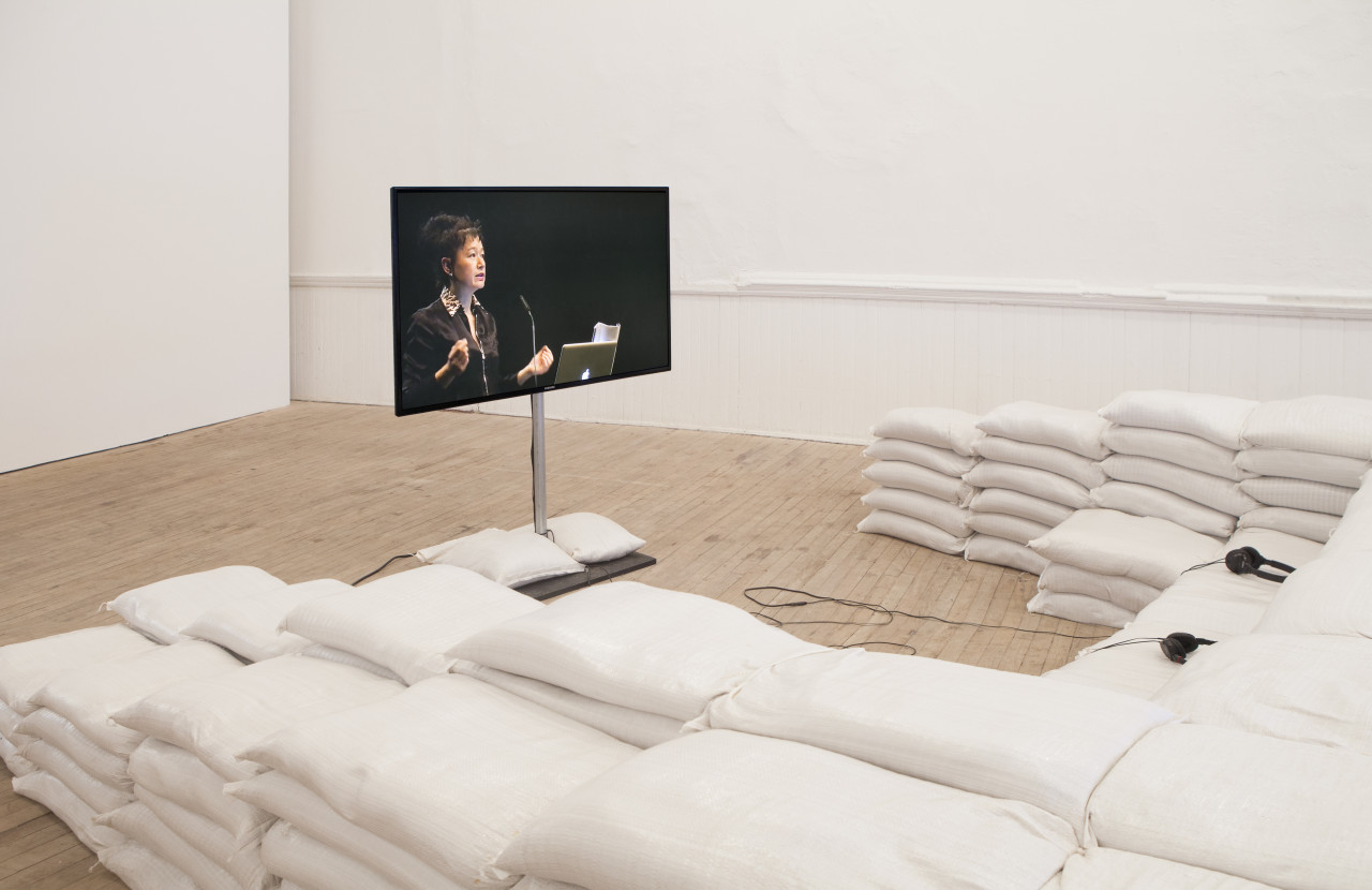 A monitor suspended in the middle of the room on a pole displays an image of Hito Steyerl speaking at a podium. Stacks of pillows, forming a seating area, surround the view of the monitor.