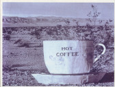 A slightly weathered sepia photograph of a sculpture of a white cup and saucer. Printed on the cup is the text 