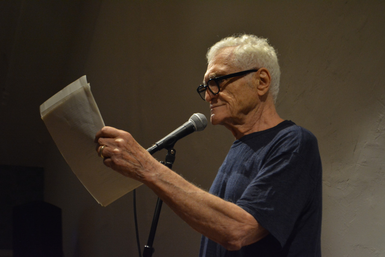 Photo of John Giorno wearing a navy blue t-shirt and black framed glasses, standing in front of a microphone and reading from loose pages held in his left hand.