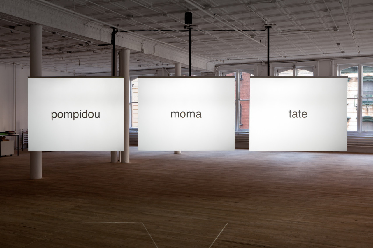 Three adjacent panels, reading "pompidou", "moma", and "tate" in black text against a luminous white background hang in a dimly-lit exhibition space.