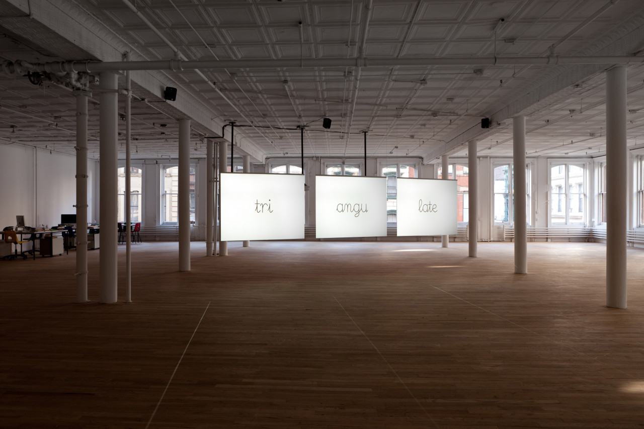In a dimly lit room with light wood flooring and white pillars, three illuminated panels are suspended from the ceiling in the distance. The panels each display a single word—"tri", "angu", and "late"—in a cursive font.