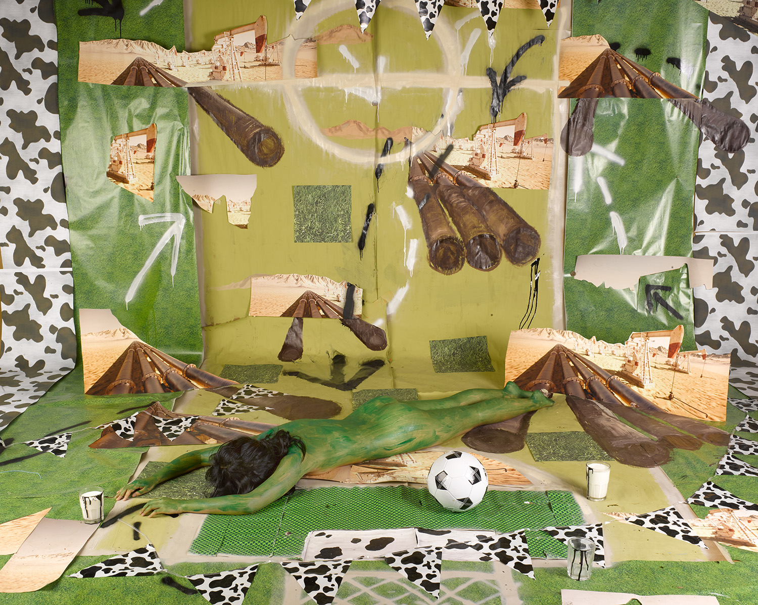 A color photograph of a performance piece showing a nude person lying face-down and completely covered in green paint. They are laying on a large set of green, patterned, and painted paper with a soccer ball and glass of white liquid near their body.