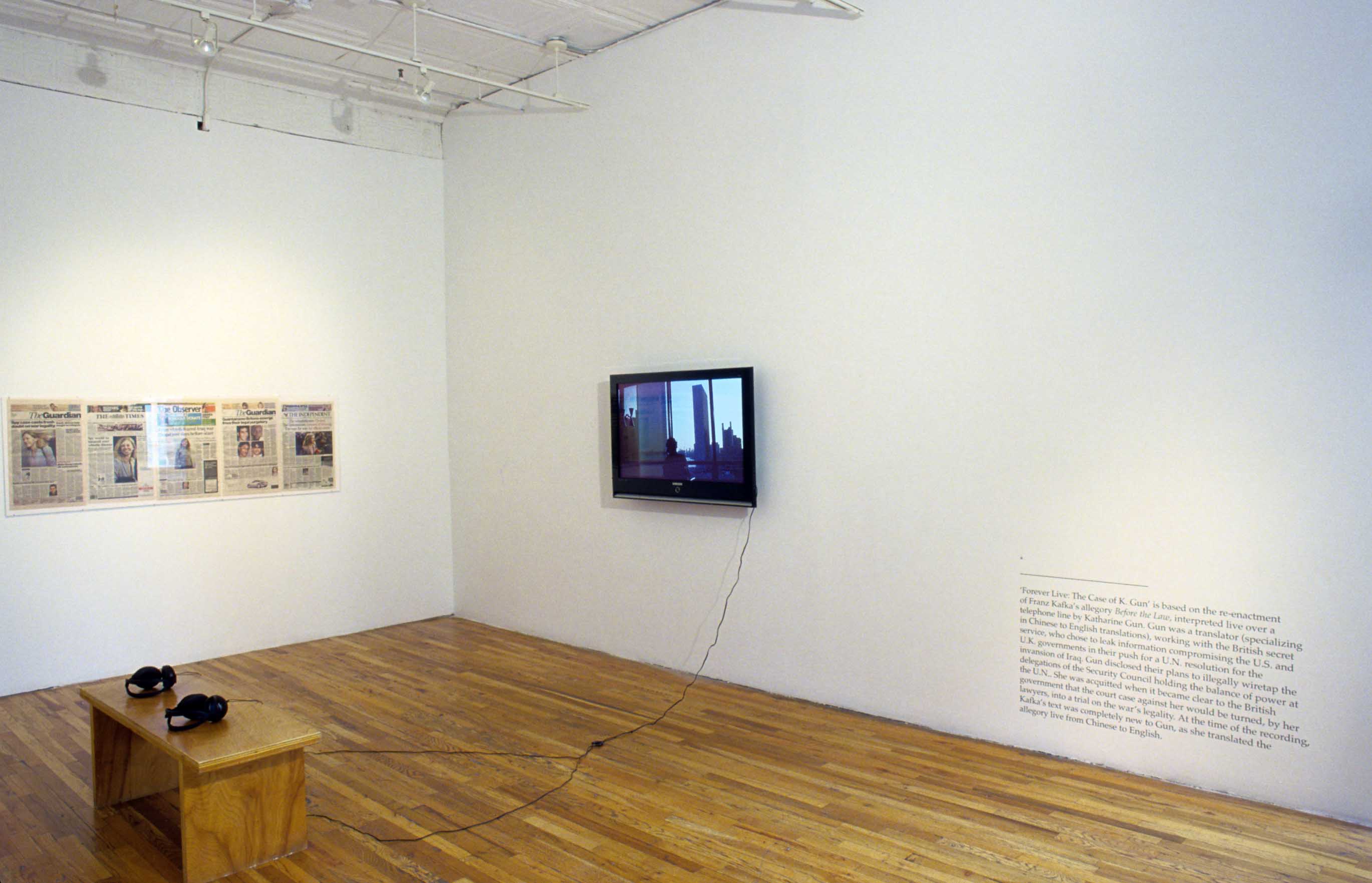 A video plays on a monitor installed on the wall of a gallery space. Two pairs of headphones attached to the monitor rest on a wooden bench facing the screen. The front pages of several newspapers are displayed on a gallery wall to the left of the monitor, while to the right, a paragraph of wall vinyl is installed on the lower half of the gallery wall.