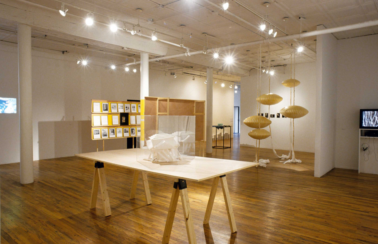 Several sculptures, video works, and models are installed in a gallery space with wooden flooring. Situated closest to the viewer is a geometric, white model installed in an acrylic box, which in turn is displayed on a large wooden table.