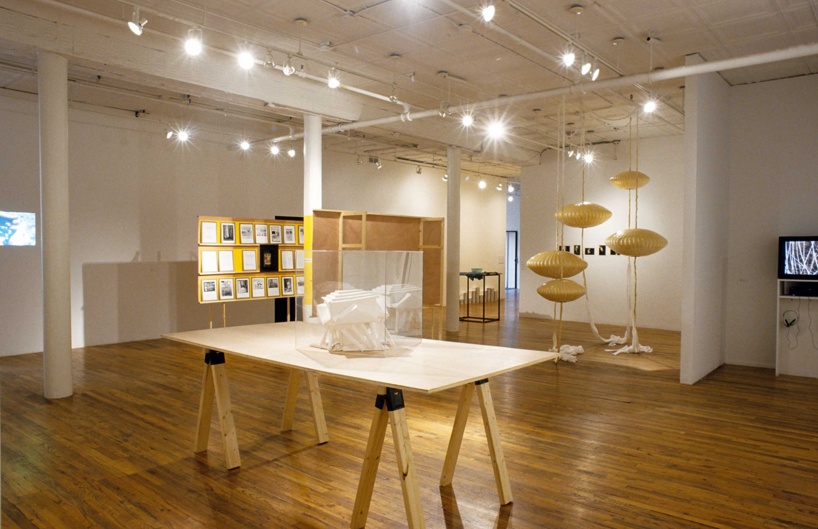 Several sculptures, video works, and models are installed in a gallery space with wooden flooring. Situated closest to the viewer is a geometric, white model installed in an acrylic box, which in turn is displayed on a large wooden table.