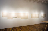 Several rectangular sheets of cream colored paper hang side-by-side on a gallery wall. The papers are arranged in clusters of three or more and appear blank, with faint creases visible on their surfaces.