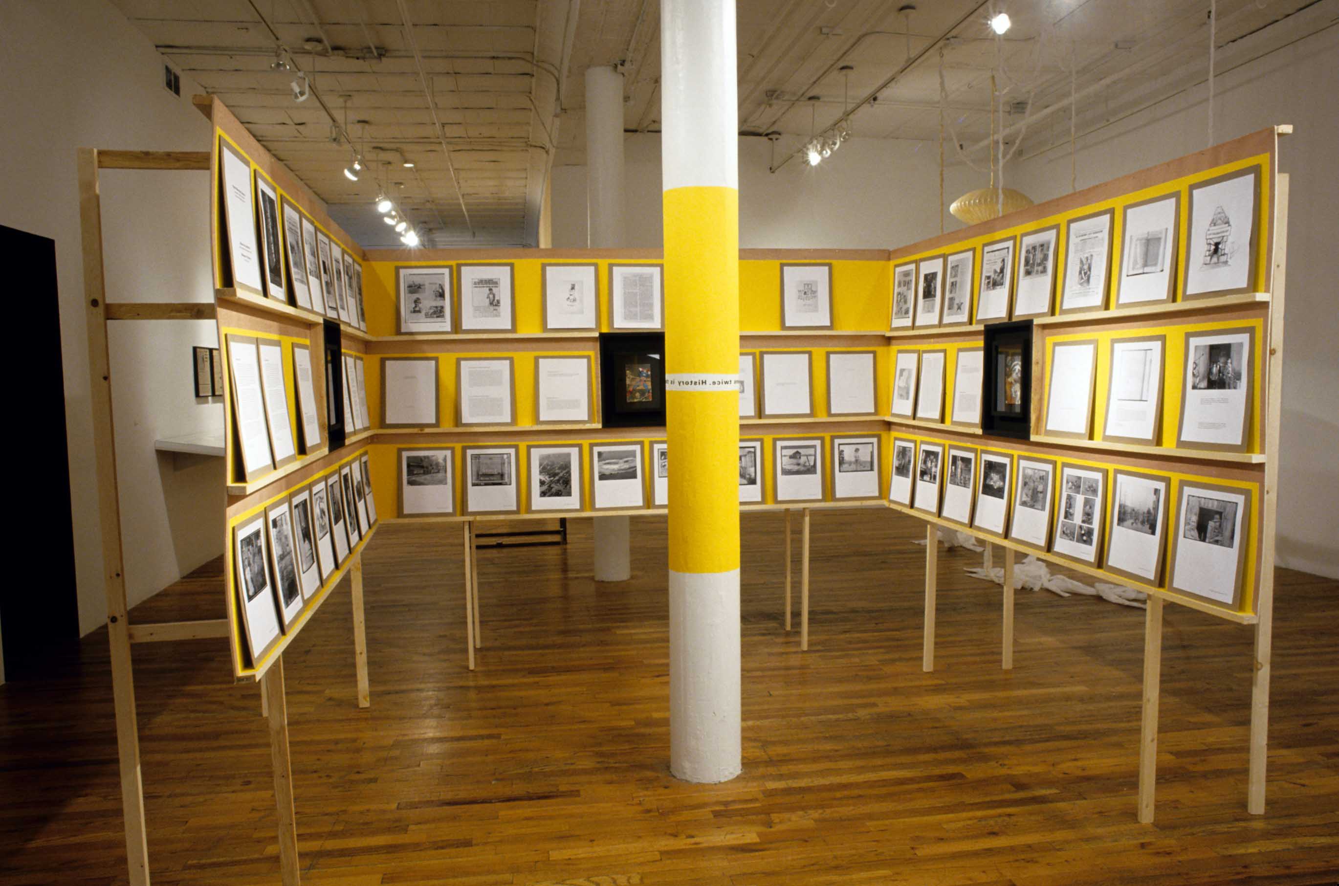 Three free-standing walls supporting a series of shelves are arranged at right angles around a pole in a gallery space. The shelves display a series of pieces of paper containing both images and text. The shelves are yellow, and a portion of the center of the column has also been painted yellow.