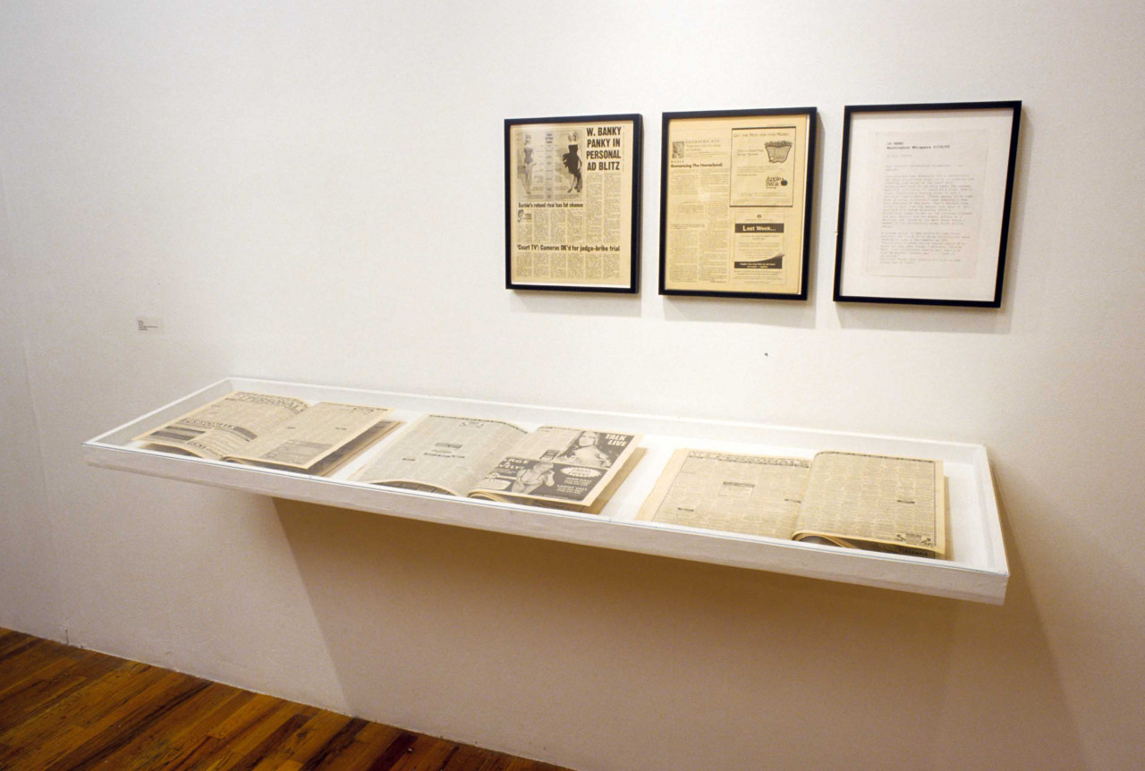 Two newspaper clippings and a sheet of written correspondence are installed in black frames on a gallery wall above a small vitrine. The vitrine displays a series of open, vintage magazines.