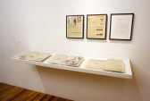 Two newspaper clippings and a sheet of written correspondence are installed in black frames on a gallery wall above a small vitrine. The vitrine displays a series of open, vintage magazines.
