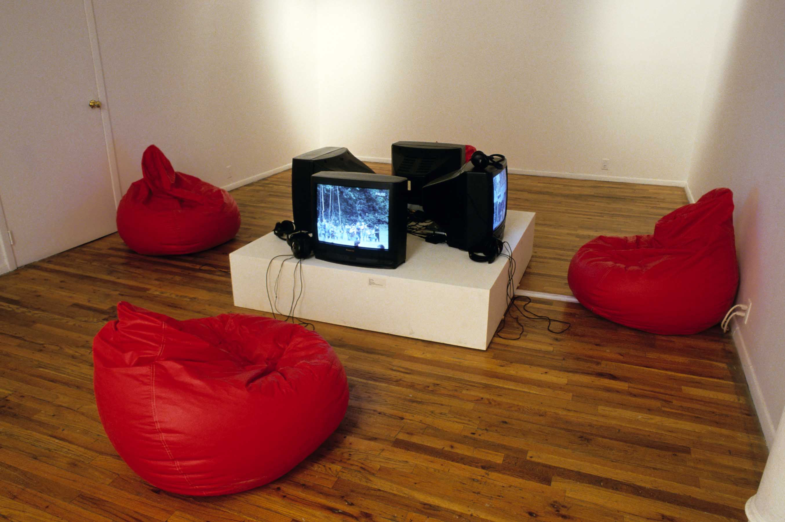 Four red bean bags are arranged on a gallery floor around four small tv monitors, each facing a different direction. The monitors are installed on a low, square plinth.