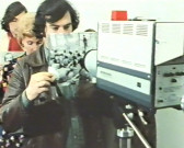 A person stands in line to hold up a black and white photograph in front of a Super 8 camera, as two people are lined up behind him.