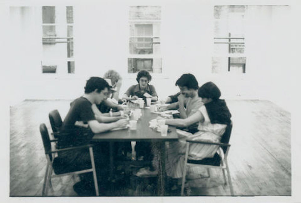 A group of people seated around a table in an empty room. In front of them are various cups and papers, they appear to be in deep thought.