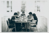 A group of people seated around a table in an empty room. In front of them are various cups and papers, they appear to be in deep thought.