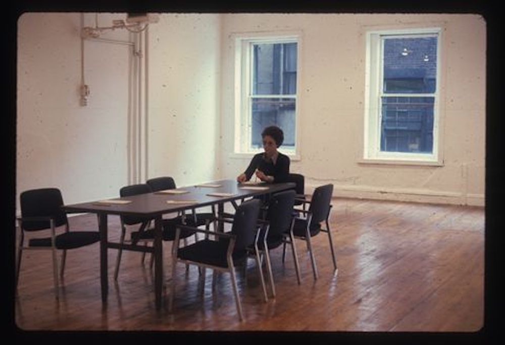 Color photograph of a figure sitting at the head of a table with seven chairs in an empty room. In front of each chair there is a small stack of papers.