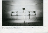 Black and white image of a studio light shining onto a set of four panels against a wall. Below, text reads 