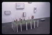 A color installation photograph of a gallery space with several framed objects hanging on the wall, and a sculpture of a green parkscape on white stilts in the middle.