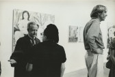 A black and white photograph of a gallery with paintinfs on the wll and several figures milling and chatting.