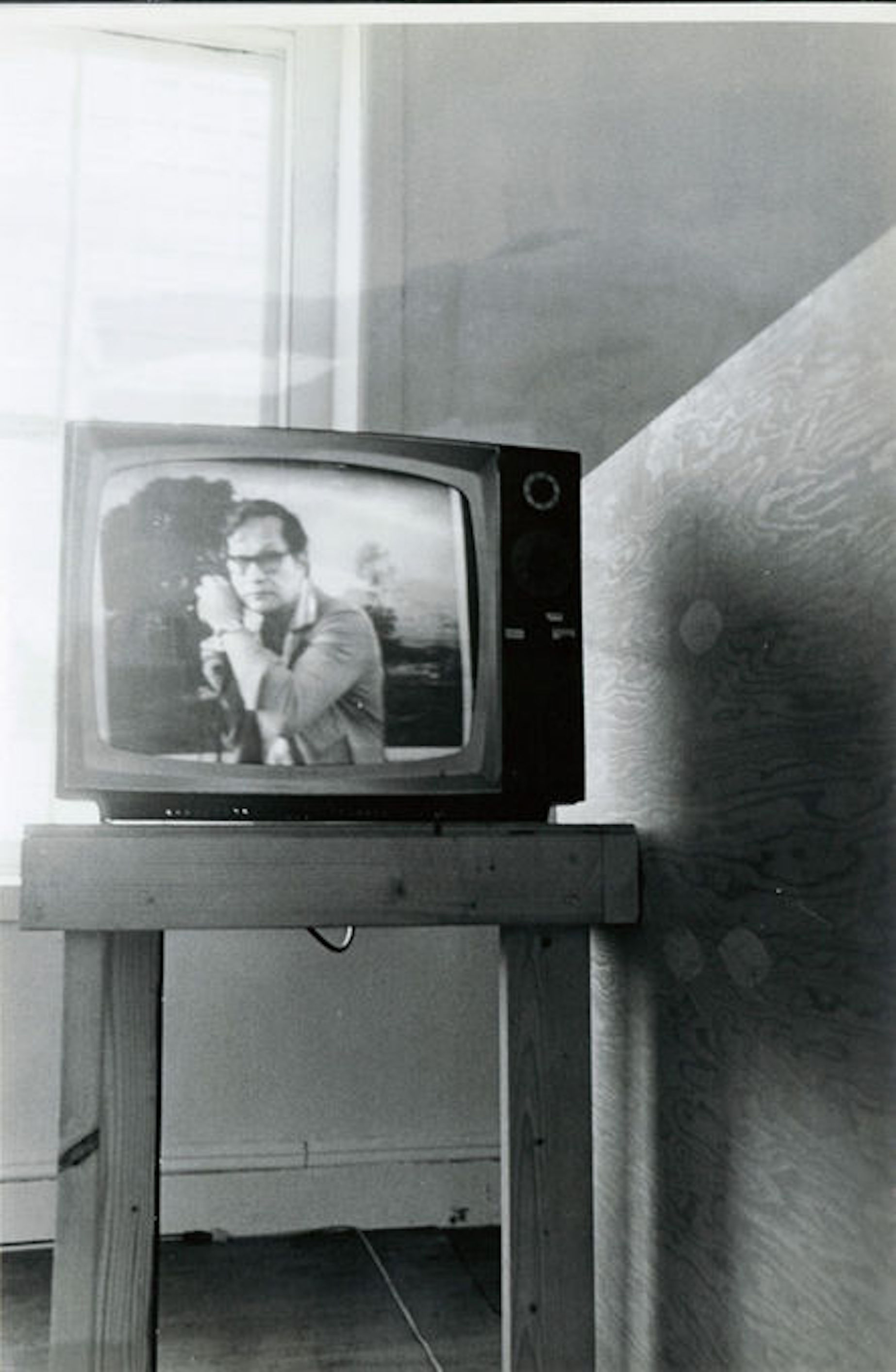 A black and white photograph of a television set upon a wooden side table in front of a window in a gallery space.