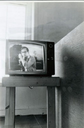 A black and white photograph of a television set upon a wooden side table in front of a window in a gallery space.