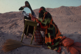 Two people in a mountainous, desert location face each other. One, dressed in an orange and green cloak, sits at a green desk. The other is posed with a mirror aimed at the clear sky.