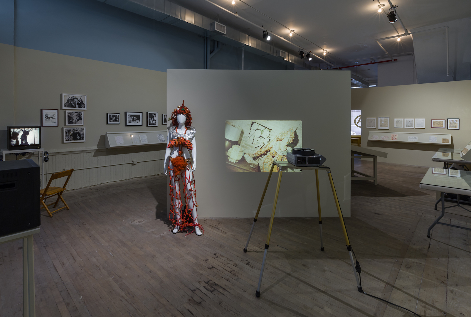A side projector projects an image of a figure in an elaborate dress onto a freestanding wall in the middle of a gallery space. A mannequin wearing a decorative orange costumes stands to the left of the projection. In the background, a video monitor sits on a table and framed documents hang on the walls.